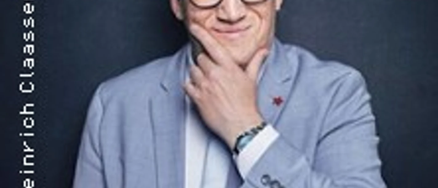 Ein Mann im blauen Sakko steht lächelnd vor einem dunklen Hintergrund, seine Hand am Kinn.