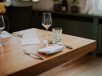 Restaurant Dreigang Herford- Lars May (3).jpg Ein stilvoll gedeckter Holztisch in einem Restaurant mit Gläsern, Besteck und einer Speisekarte.