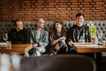 Vier Personen sitzen lächelnd in einem Restaurant an einem Holztisch vor einer roten Backsteinwand.