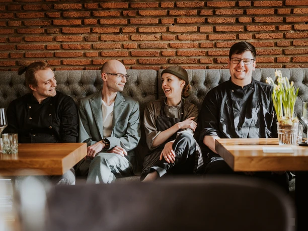 Vier Personen sitzen lächelnd in einem Restaurant an einem Holztisch vor einer roten Backsteinwand.