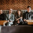 Restaurant Dreigang Herford- Lars May (2).jpg Vier Personen sitzen lächelnd in einem Restaurant an einem Holztisch vor einer roten Backsteinwand.