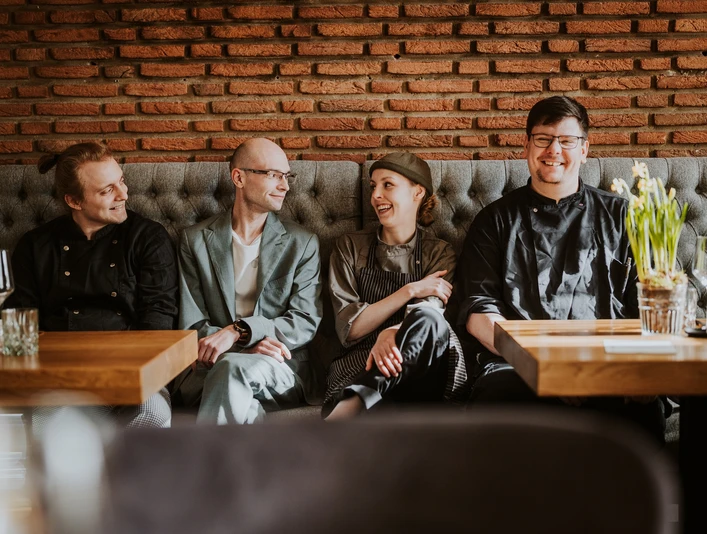 Restaurant Dreigang Herford- Lars May (2).jpg Vier Personen sitzen lächelnd in einem Restaurant an einem Holztisch vor einer roten Backsteinwand.