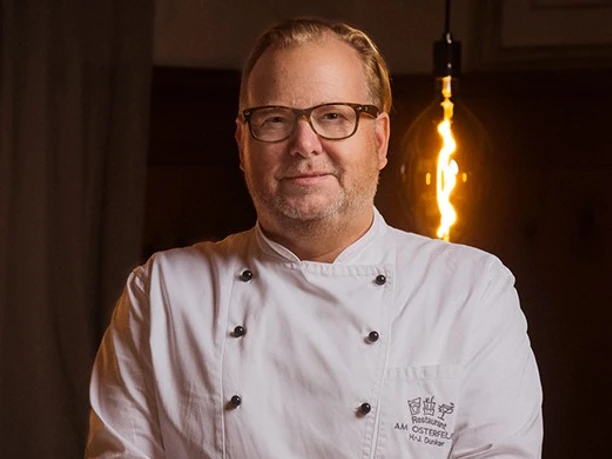 am-osterfeuer-2.jpg Porträt eines Kochs im weißen Kittel, vor dunklem Hintergrund mit warmem Licht.