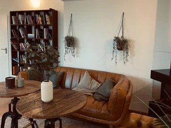 Sauna im Denkwerk Herford_Aufenthaltsbereich 2.jpg Moderner Aufenthaltsbereich mit Ledersofa, Bücherregal und hängenden Pflanzen an weißen Wänden.