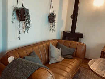 Sauna im Denkwerk Herford_Aufenthaltsbereich 1.jpg Ein einladendes Ledersofa mit Kissen und Decke, umgeben von hängenden Pflanzen und einem Kamin.