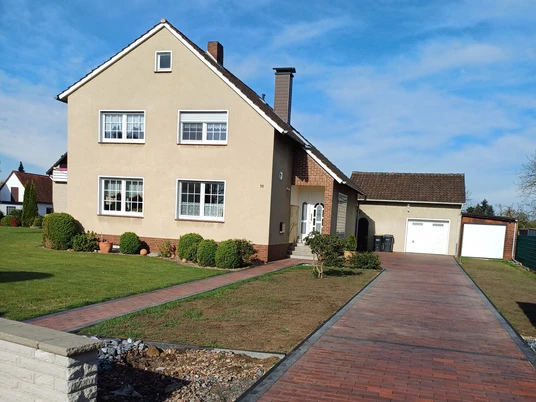 Zimmervermietung Busse Zweistöckiges Haus mit hellem Anstrich, gepflegtem Vorgarten und gepflasterter Einfahrt.