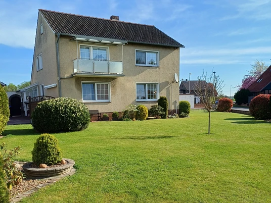 Zimmervermietung Busse Zweistöckiges Wohnhaus mit gepflegtem Garten, Sträuchern und wenig Bewölkung im Hintergrund.