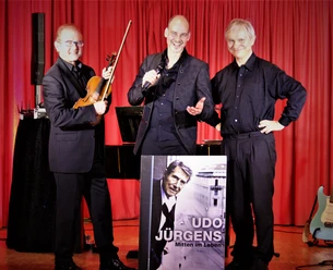 Trio.JPG Drei Musiker stehen lächelnd vor einem roten Vorhang; einer hält eine Violine, ein anderer ein Mikrofon, und ein dritter mit verschränkten Armen präsentieren ein Plakat von Udo Jürgens.