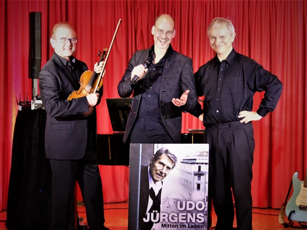 Trio.JPG Drei Musiker stehen lächelnd vor einem roten Vorhang; einer hält eine Violine, ein anderer ein Mikrofon, und ein dritter mit verschränkten Armen präsentieren ein Plakat von Udo Jürgens.