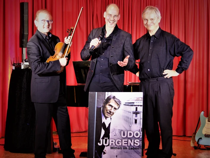 Trio.JPG Drei Musiker stehen lächelnd vor einem roten Vorhang; einer hält eine Violine, ein anderer ein Mikrofon, und ein dritter mit verschränkten Armen präsentieren ein Plakat von Udo Jürgens.