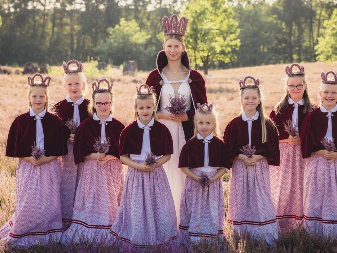 heidebluetenfest-amelinghausen-erlebniscard.jpg Eine Gruppe von Mädchen in traditionellen Dirndln und Kronen steht lächelnd in der blühenden Heide.