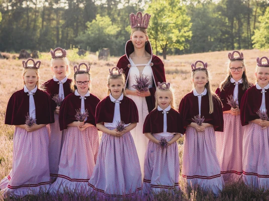 heidebluetenfest-amelinghausen-erlebniscard.jpg Eine Gruppe von Mädchen in traditionellen Dirndln und Kronen steht lächelnd in der blühenden Heide.