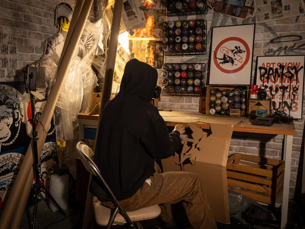 Banksy bei der Arbeit - House of Banksy Leipzig In der Ausstellung des House of Banksy in Leipzig steht eine Werkbank mit Spraydosen, an der eine vermummte Puppe sitzt, die den unbekannten Künstler Banksy bei der Arbeit darstellen soll