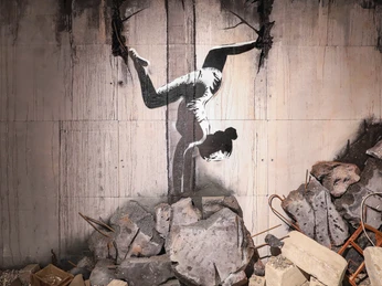 Turnerin in der Ukraine - House of Banksy Leipzig Bild aus der Ausstellung im House of Banksy in Leipzig von einem Graffiti einer Turnerin, die einen Handstand macht, das der Künstler in den Trümmern in der Ukraine gesprüht hat