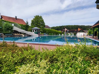 Waldfreibad Preußisch Oldendorf mit großer Wasserrutsche, umgeben von grüner Natur und blauen Himmel.