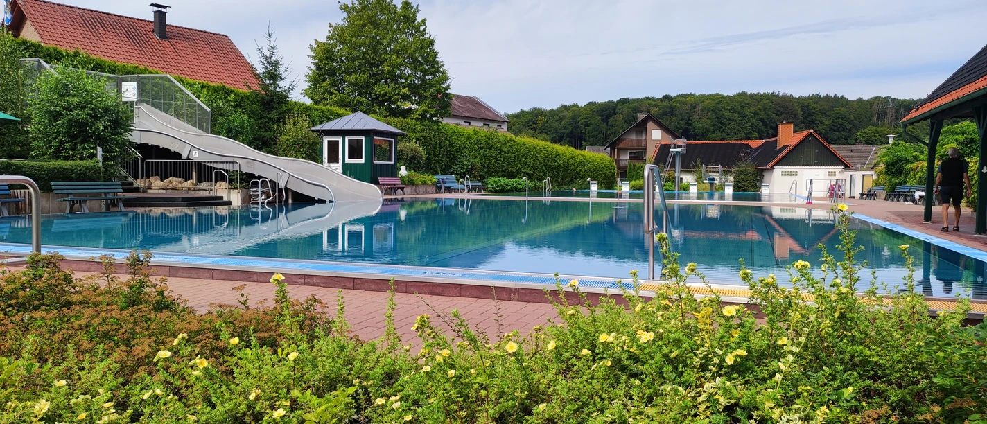 Waldfreibad Preußisch Oldendorf mit großer Wasserrutsche, umgeben von grüner Natur und blauen Himmel.