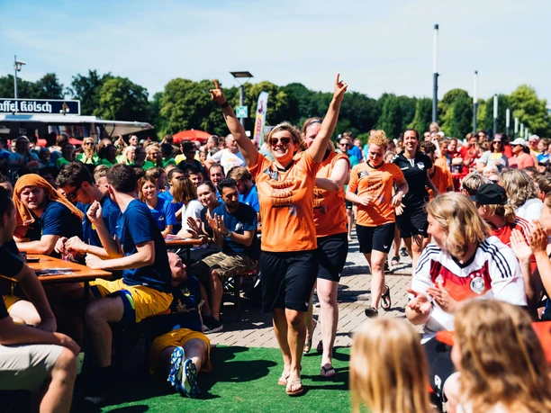 A cheering crowd watches the Come Together Cup in Cologne in sunny weather. The foreground shows people in colourful jerseys, cheering and clapping. An atmosphere full of joy and community.