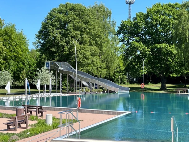 Freibad mit großer Wasserrutsche im Grünen, umgeben von Bäumen und klarblauem Himmel.