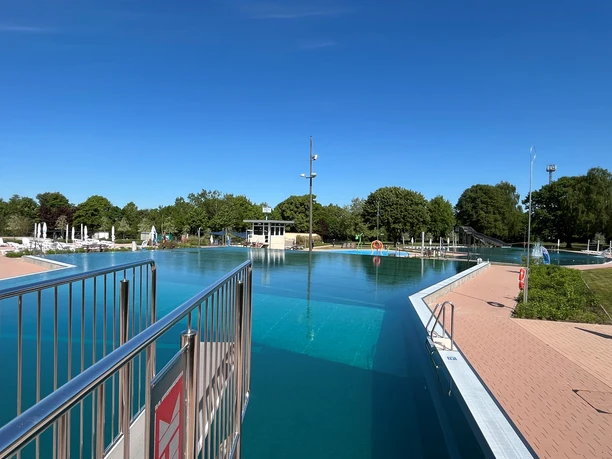 A spacious outdoor pool with crystal-clear water, surrounded by green trees and a blue sky.