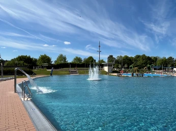 Blaues Freibadbecken mit Fontäne, umgeben von Bäumen und blauem Himmel, lädt zum Schwimmen ein.