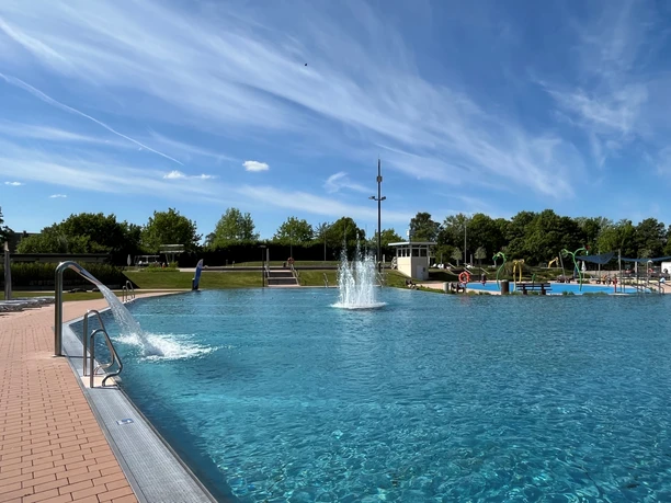 Blaues Freibadbecken mit Fontäne, umgeben von Bäumen und blauem Himmel, lädt zum Schwimmen ein.