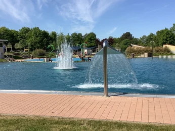 Ein modernes Freibad mit sprudelnden Wasserspielen, umgeben von grünen Bäumen bei strahlendem Sonnenschein.