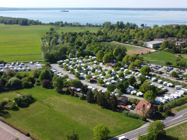 Wohnmobilstellplatz Luftbild Luftaufnahme eines großen Wohnmobilstellplatzes, umgeben von grünen Feldern und einem See im Hintergrund.