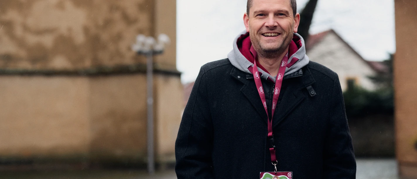 Stadtführer Stefan Jülke.jpg Ein lächelnder Mann steht in einem Innenhof vor einer historischen Gebäudefassade. Er trägt einen schwarzen Mantel und hat eine rote Umhängeleine um den Hals. Die Umgebung wirkt ruhig und fachwerkartig, mit vereinzelt stehenden Laternen und kahlen Bäumen im Hintergrund. Ein einladender Tag für eine geführte Stadterkundung.