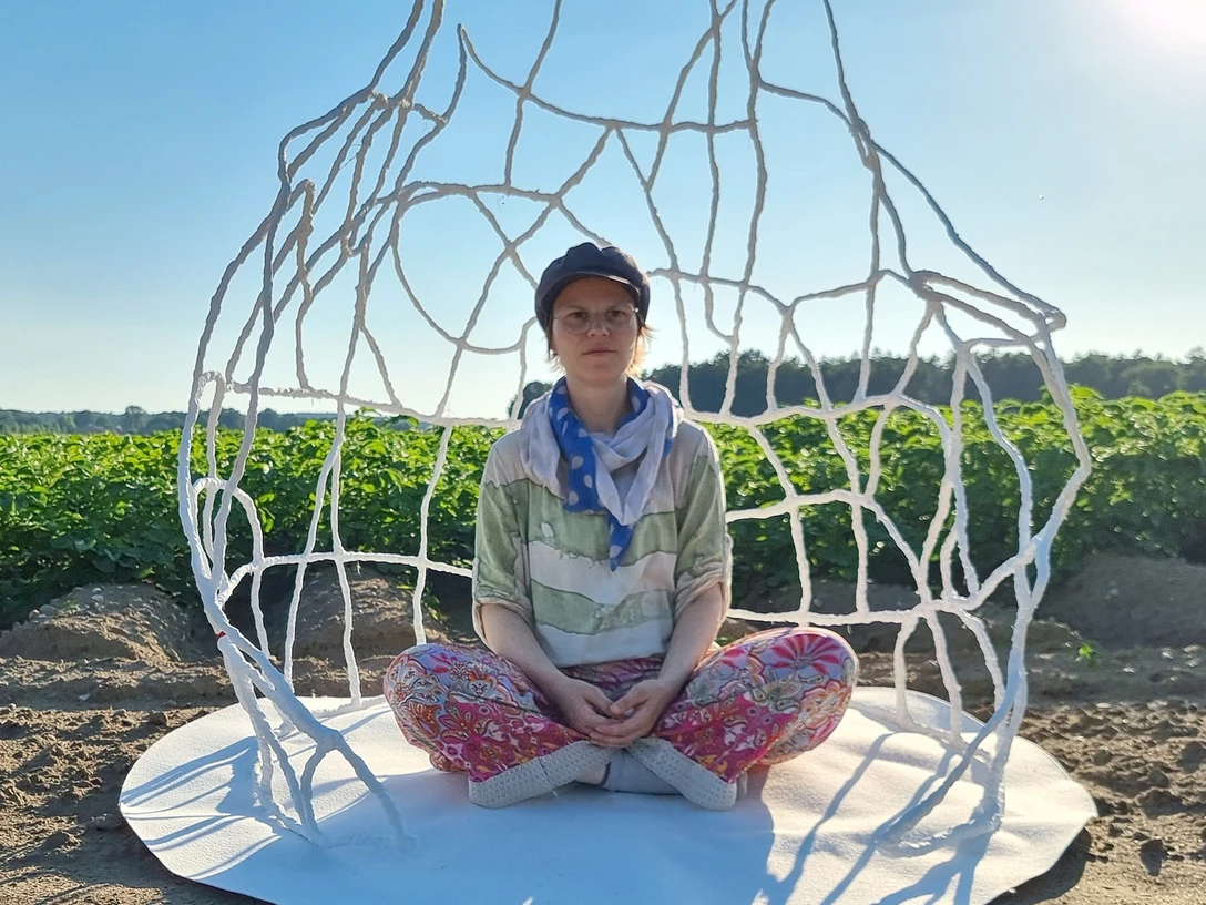 kuenstlerin-im-raumkoerper-jessca-kulp-2025.jpg Eine Künstlerin sitzt meditativ in einem filigranen, weißen Drahtkunstwerk auf einem Feld unter blauem Himmel.