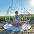 kuenstlerin-im-raumkoerper-jessca-kulp-2025.jpg Eine Künstlerin sitzt meditativ in einem filigranen, weißen Drahtkunstwerk auf einem Feld unter blauem Himmel.