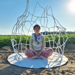 kuenstlerin-im-raumkoerper-jessca-kulp-2025 Eine Künstlerin sitzt meditativ in einem filigranen, weißen Drahtkunstwerk auf einem Feld unter blauem Himmel.