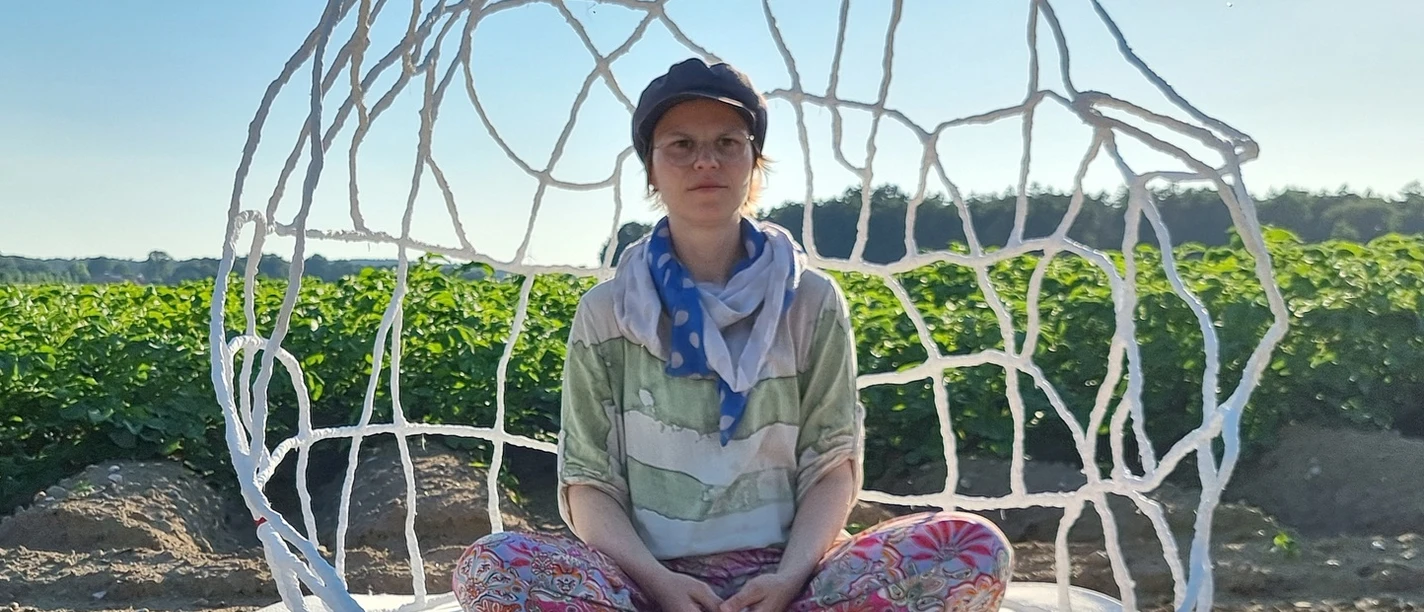 kuenstlerin-im-raumkoerper-jessca-kulp-2025 Eine Künstlerin sitzt meditativ in einem filigranen, weißen Drahtkunstwerk auf einem Feld unter blauem Himmel.