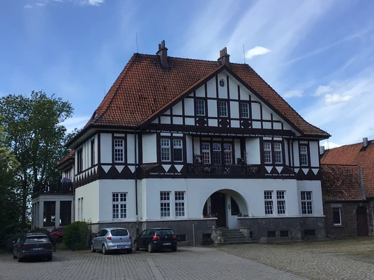Ein traditionelles Fachwerkhaus mit roten Ziegeln, umgeben von geparkten Autos bei sonnigem Wetter.A traditional half-timbered house with red bricks, surrounded by parked cars in sunny weather.Et traditionelt bindingsværkshus med røde mursten, omgivet af parkerede biler i solskinsvejr.Een traditioneel vakwerkhuis met rode bakstenen, omringd door geparkeerde auto's bij zonnig weer.