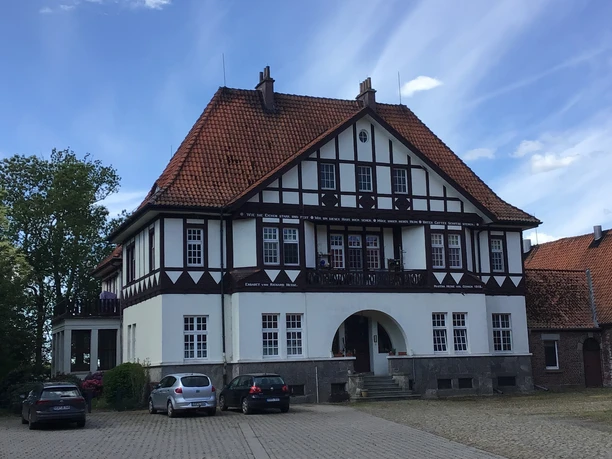 A traditional half-timbered house with red bricks, surrounded by parked cars in sunny weather.