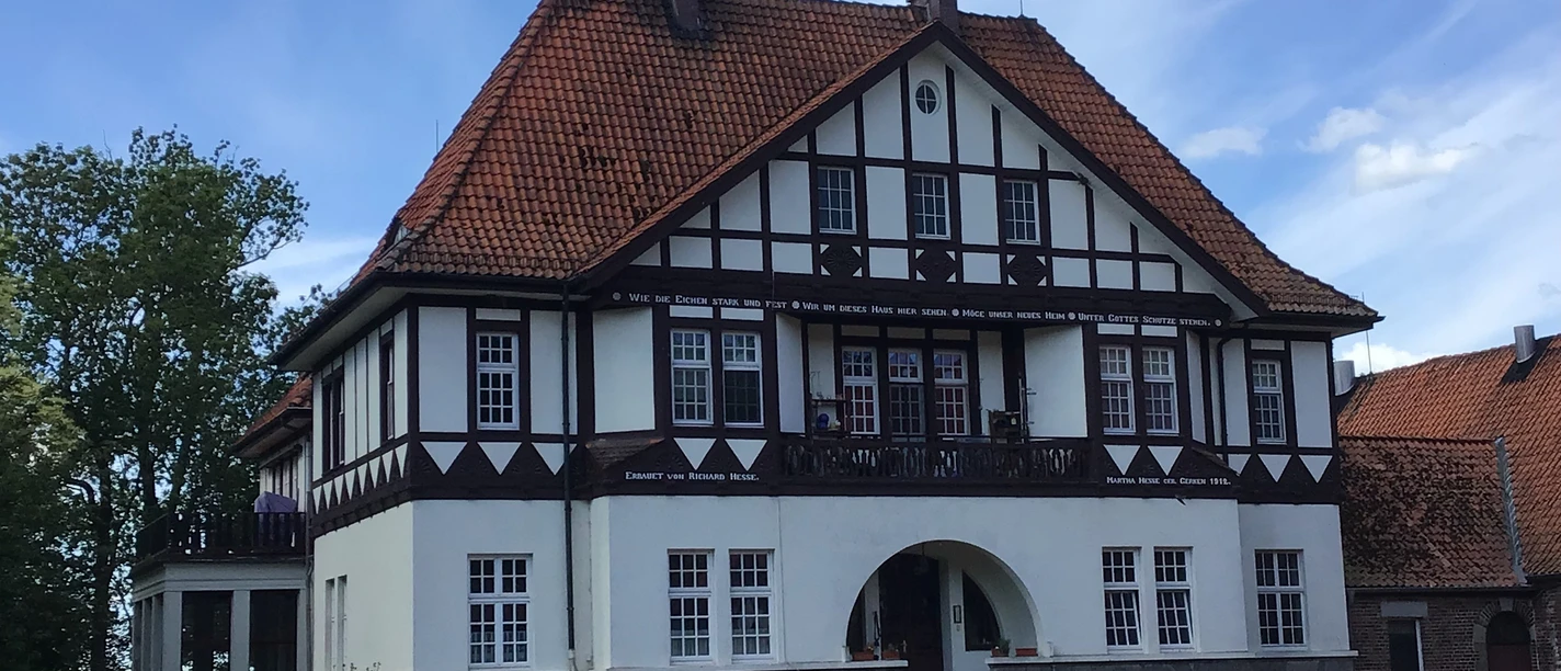 FeWo Gut Lohof Ein traditionelles Fachwerkhaus mit roten Ziegeln, umgeben von geparkten Autos bei sonnigem Wetter.
