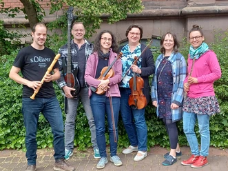 20250508_133614.jpg Eine fröhliche Musikgruppe mit sechs Personen, die verschiedene Instrumente vor einer grünen Hecke spielen.
