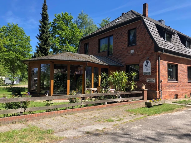 Fachwerkhaus Bruchhausen Ein rotes Backsteinhaus mit Satteldach, Wintergarten, umgeben von grünen Bäumen an einem sonnigen Tag.