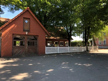 Brick building with riding stable and white fence, surrounded by deciduous trees under a bright blue sky.