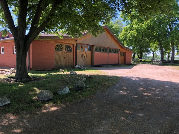 A riding stable with a red brick building and a wooden façade, surrounded by large, shady trees.