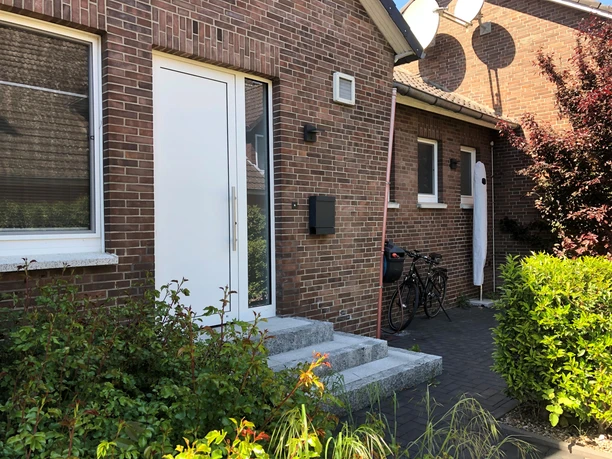 Two bicycles are parked in front of the front door of a brick house with small plants in the foreground.
