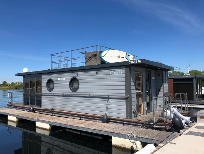 Hausboot mit modernen, grauen Fassaden, auf ruhigem Wasser im Hafen, Sonnenterrasse mit Surfbrettern.Houseboat with modern, gray facades, on calm water in the harbor, sun terrace with surfboards.Husbåd med moderne, grå facader, på roligt vand i havnen, solterrasse med surfbrætter.Woonboot met moderne, grijze gevels, op rustig water in de haven, zonneterras met surfplanken.