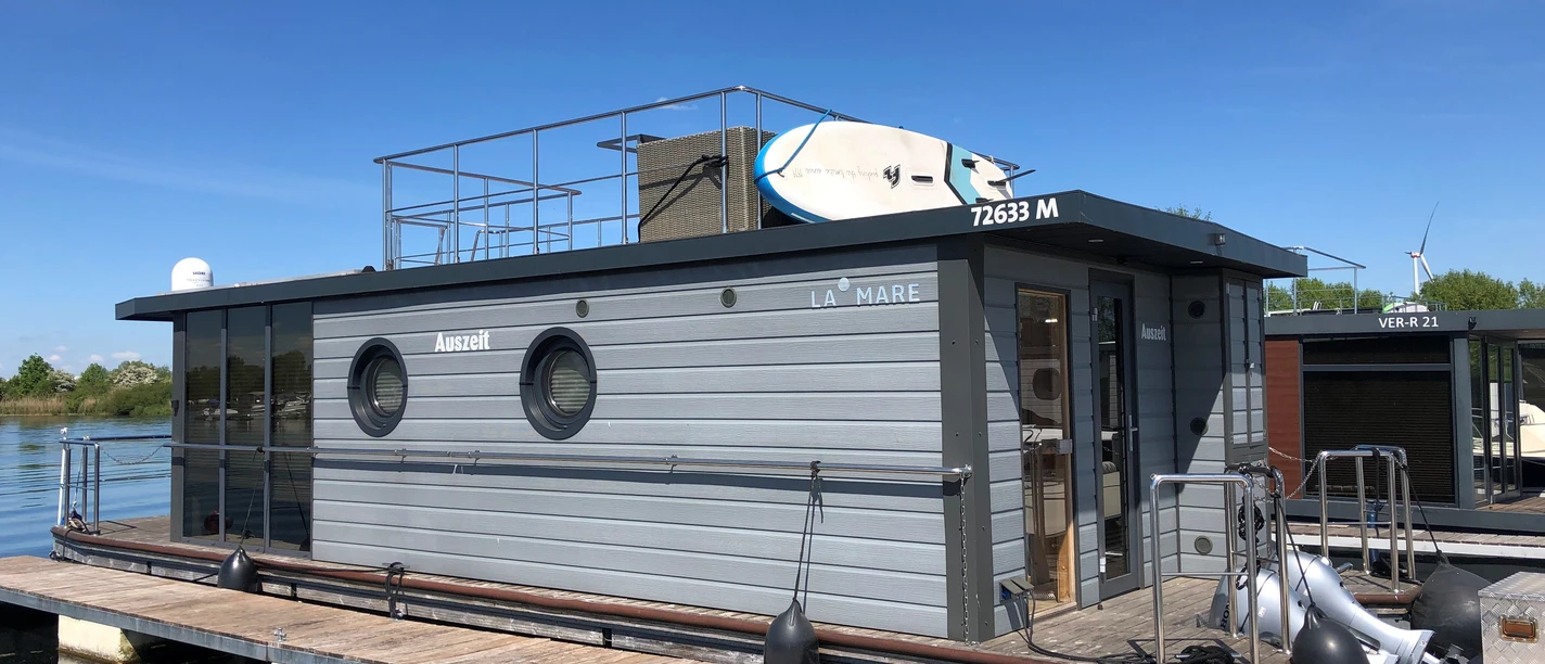 La Mare Hausboot Wieltsee Houseboat with modern, gray facades, on calm water in the harbor, sun terrace with surfboards.