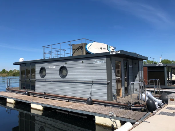 La Mare Hausboot Wieltsee Hausboot mit modernen, grauen Fassaden, auf ruhigem Wasser im Hafen, Sonnenterrasse mit Surfbrettern.