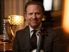 Ein lächelnder Violinist in formeller Kleidung sitzt in einem gedämpft beleuchteten Raum mit Geige.