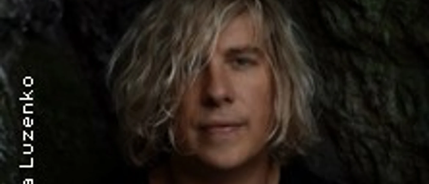 Ein Mann mit schulterlangem blonden Haar steht entspannt vor einem dunklen, natürlichen Hintergrund.