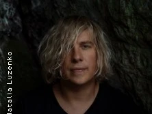 Ein Mann mit schulterlangem blonden Haar steht entspannt vor einem dunklen, natürlichen Hintergrund.