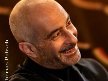 Ein Mann mit Glatze und Bart lächelt seitlich. Er trägt einen dunklen Pullover und sitzt entspannt.