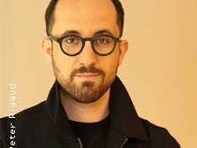 Ein Mann mit dunklem Haar und Bart trägt eine Brille. Der Hintergrund ist in warmen Tönen gehalten.