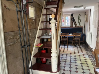 Innenansichten Museumscafé Gütersloh Rustikale Holztreppe in einem gemütlichen Raum mit Tisch und Stühlen auf gefliestem Boden.