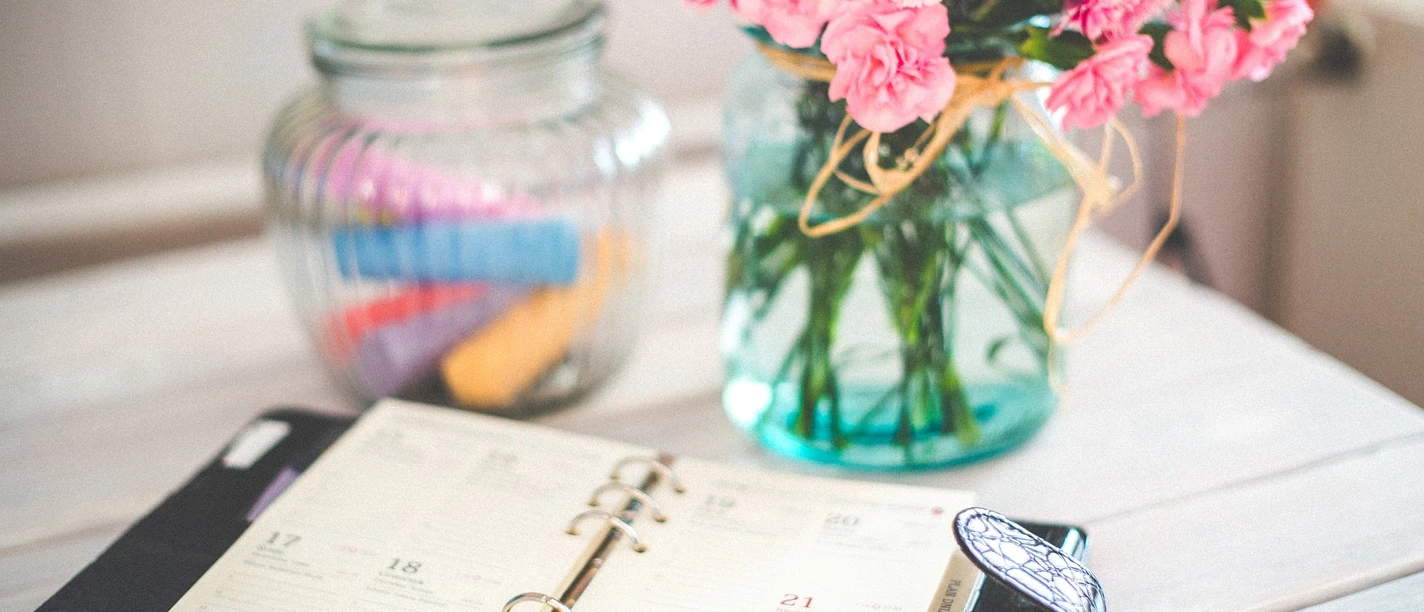 Bullet Journal Ein geöffneter Planer liegt auf einem Tisch neben einem Kerzenglas und einem Glas mit rosa Blumen.